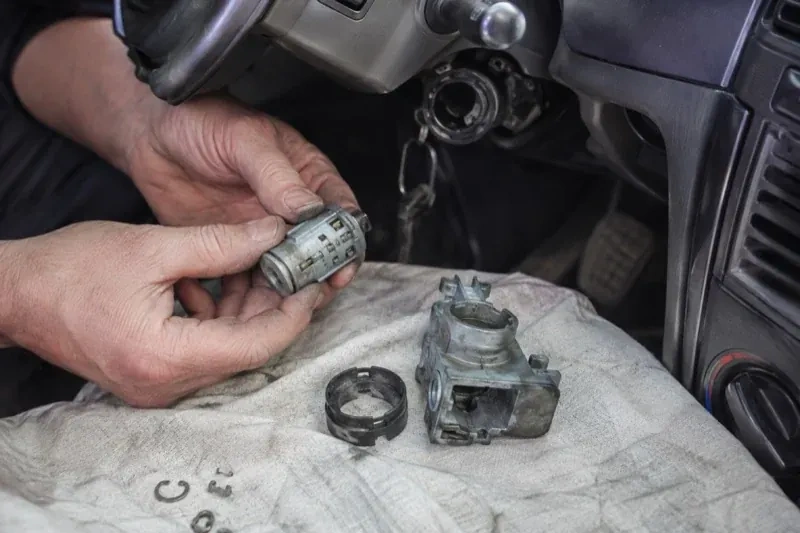 Hands holding a removed ignition cylinder beside a car steering column, with the housing and small parts on a cloth. — Ignition Lock Repair, Markham
