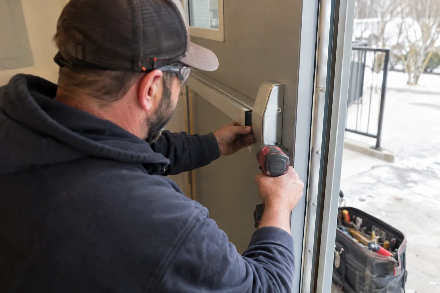 Technician in a cap and safety glasses holding a push bar end cap against a metal door with a red drill. — Markham